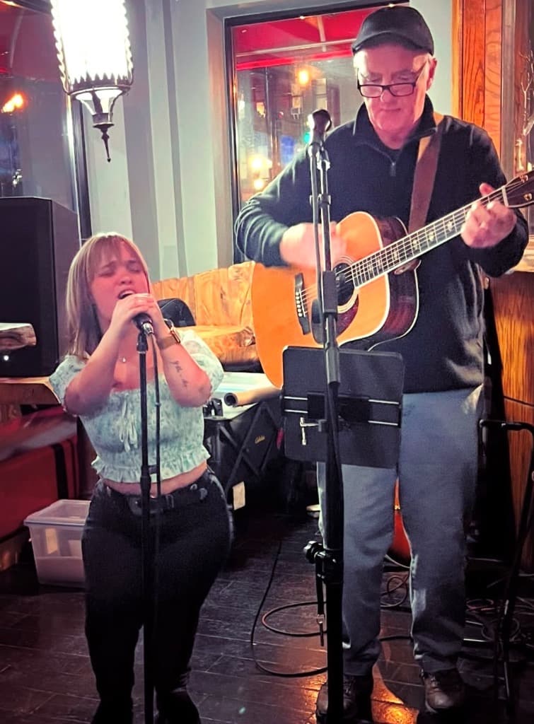 Lyndsey singing and Zach playing Martin guitar under warm dining room lights at Hamilton Restaurant